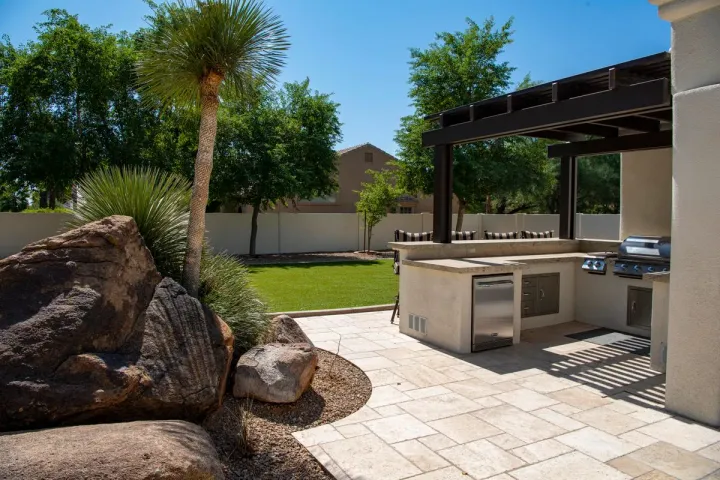 An outdoor kitchen island with grill, refrigerator, and bar seating beneath a timber pergola