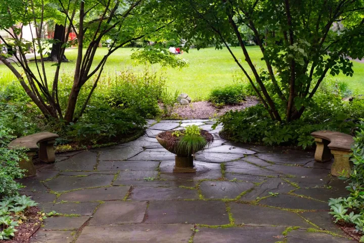 A shaded natural stone patio with mossy joints and garden benches under mature trees